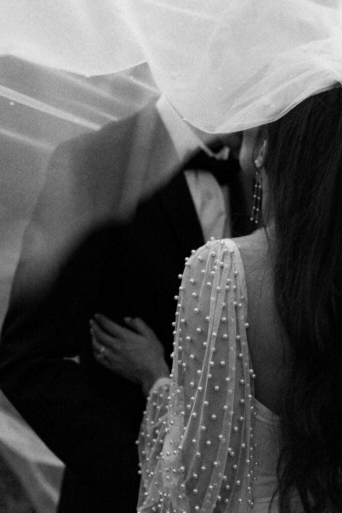 Bride wearing pearls and groom wearing a tux kissing under a veil